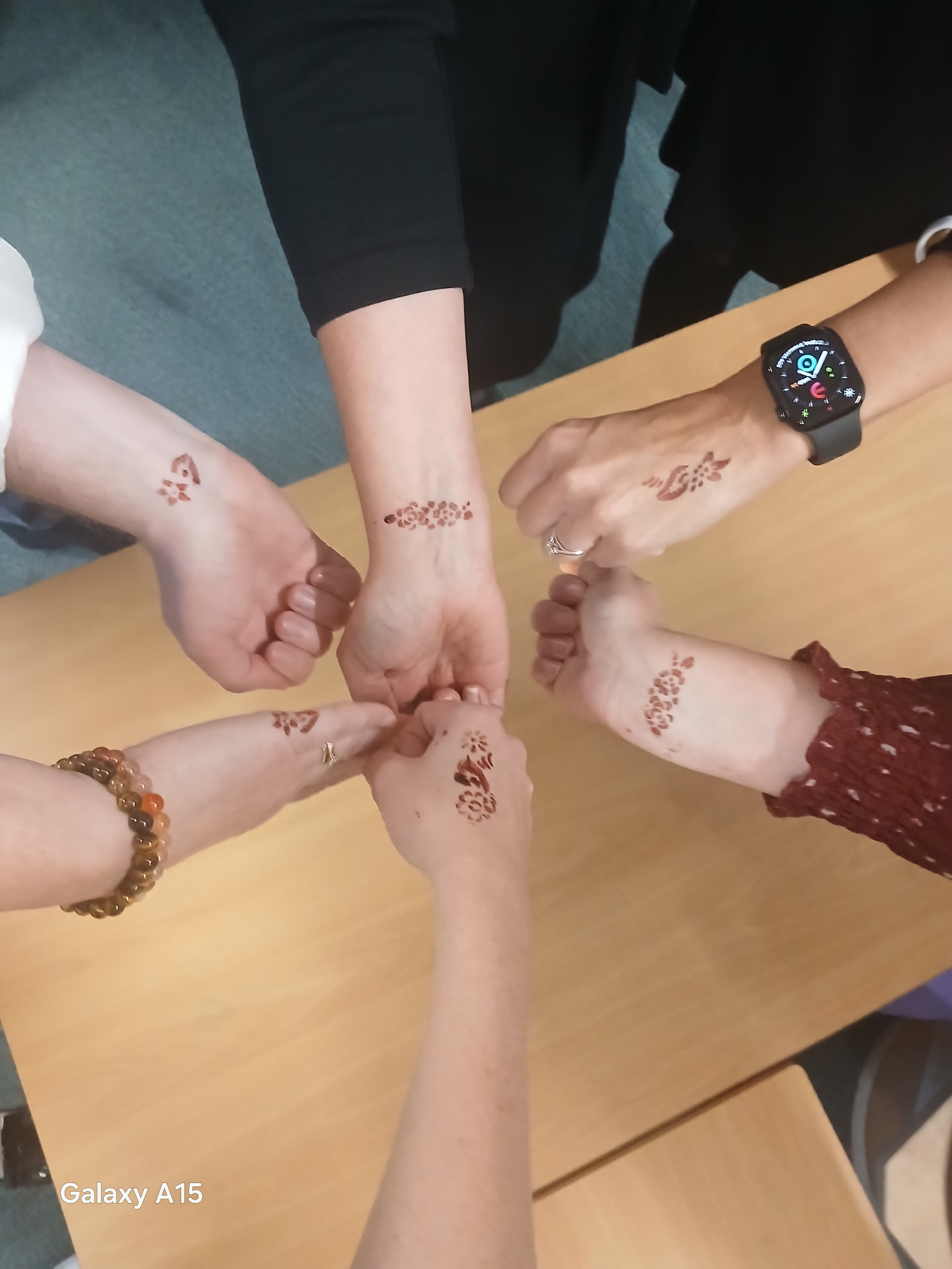 Six wrists arranged in a circle on a table, each with a small floral henna design.