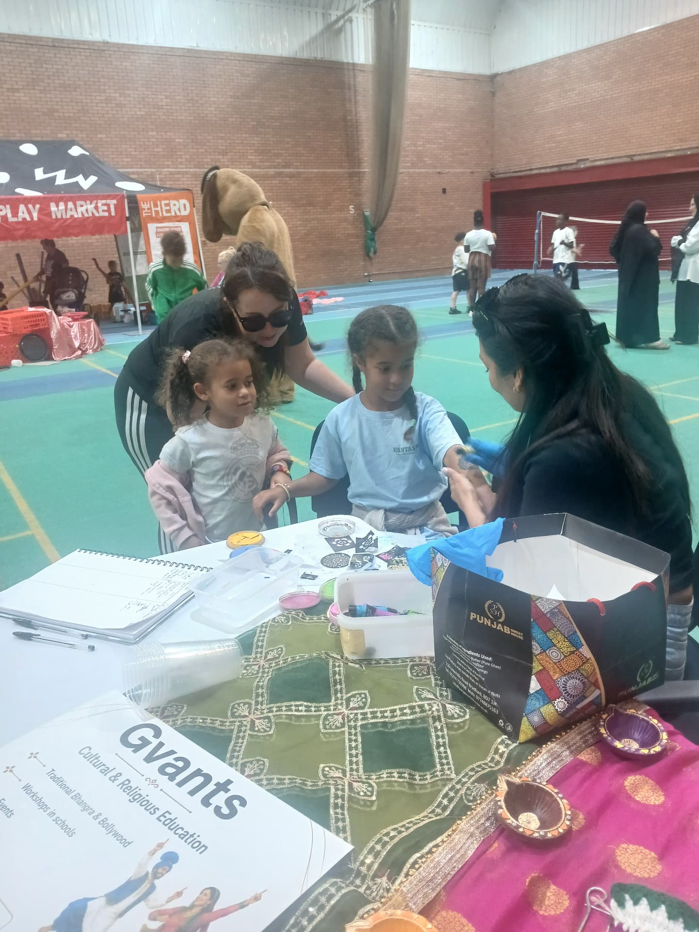 A Gvants cultural education stall at a community event, with children having henna applied and diva lamps and fabrics on the table.