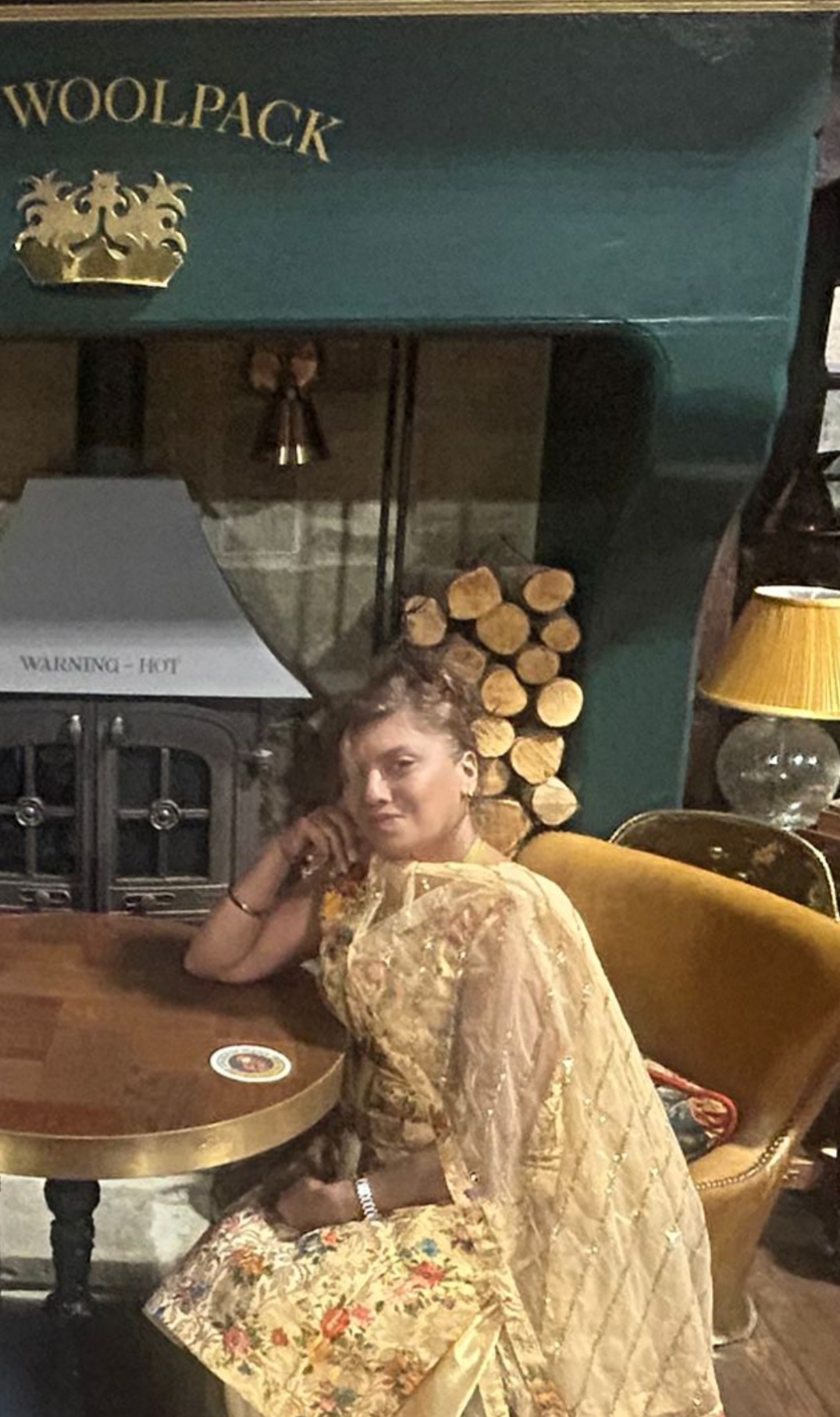 A person in a gold embroidered outfit seated at a table beneath a Woolpack sign.