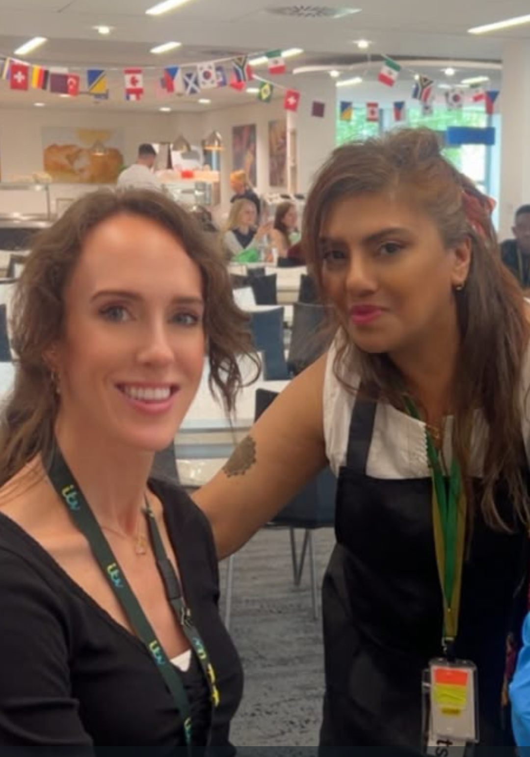 Two colleagues at an office event with international flag bunting, one showing a fresh henna design on her arm.