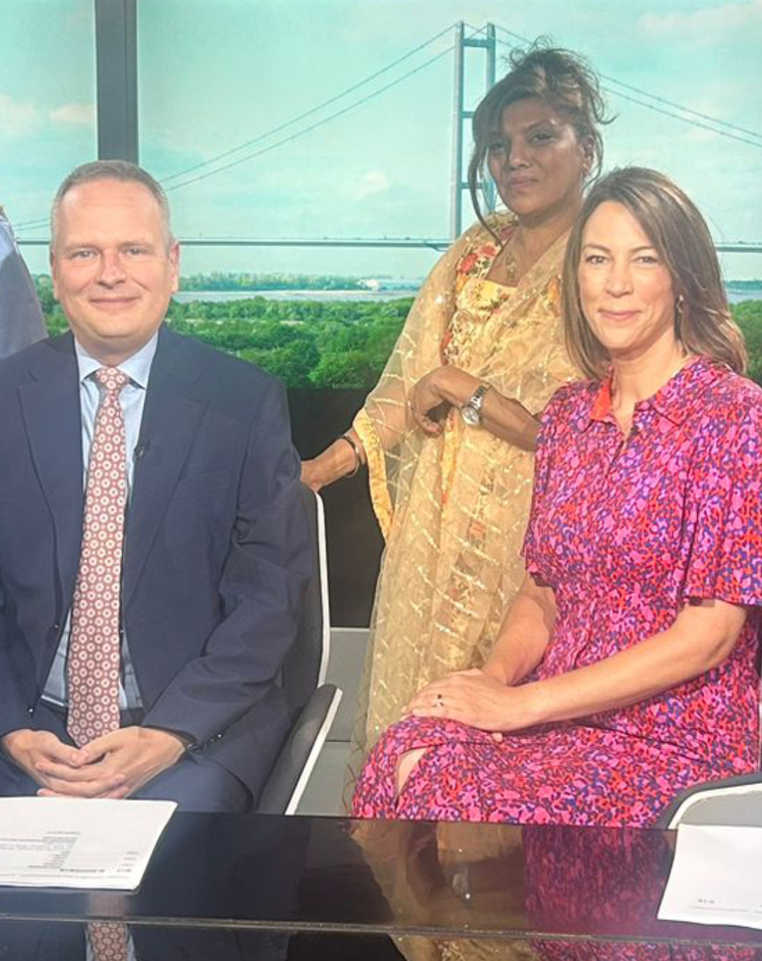A person in a gold saree standing behind two seated colleagues at a studio desk with a suspension bridge backdrop.