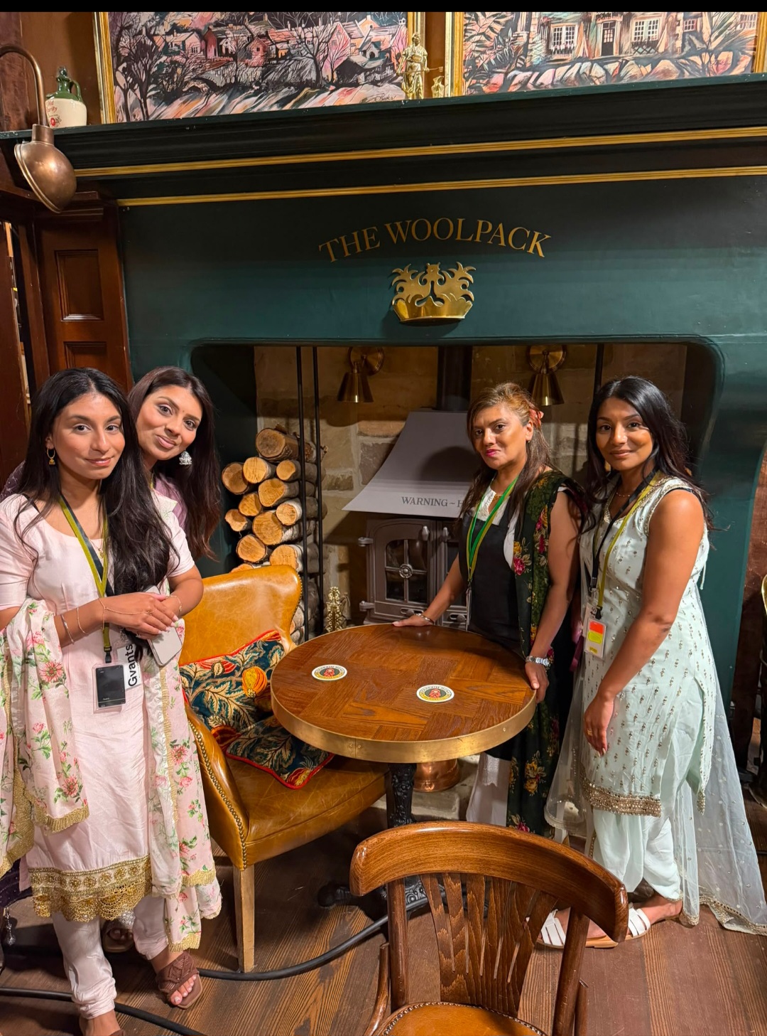 Four people in traditional South Asian outfits standing beside a fireplace under a painted Woolpack sign.