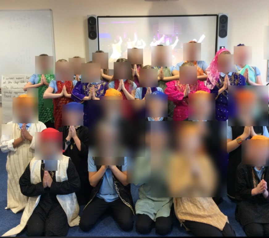 A class of pupils wearing colourful South Asian clothing and turbans, posing with hands together in a classroom. Faces are blurred.